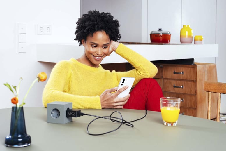 Une jeune femme est assise à une table avec un chargeur rapide Samsung et sourit en direction de son smartphone.