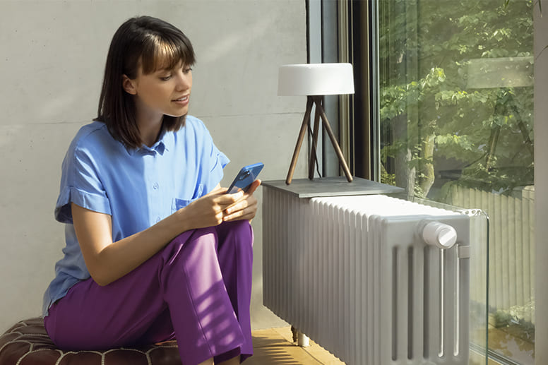 Une personne regarde un téléphone portable et se trouve devant un radiateur à thermostat intelligent.