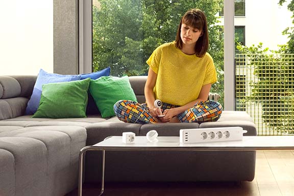 Une personne est heureuse à la table de son salon. Devant elle se trouvent différents appareils Smart Home autorisés pour les logements en location.